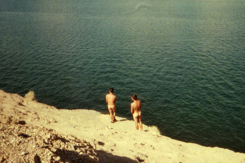 two nude people preparing to recreationally jump from a cliff into a lake