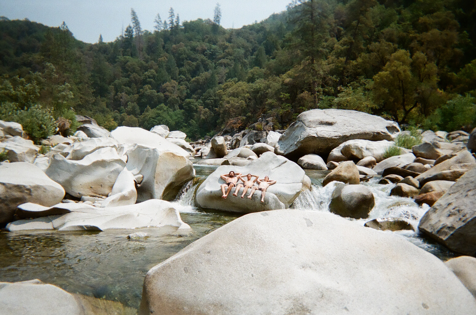 three people in swimwear lean against a rock posing with their hands behind their head